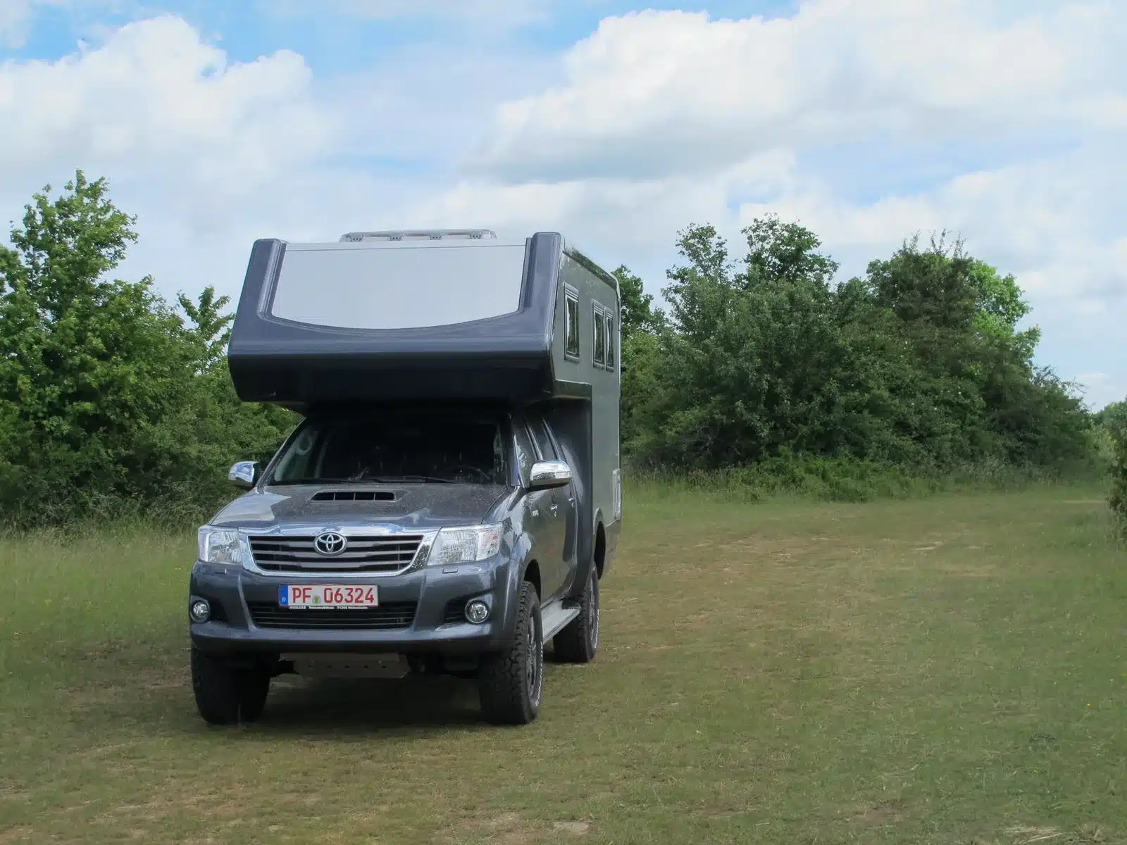 Ein Autark Hilux Offroad-Camper steht frei auf einer Wiese, umgeben von Bäumen und blauem Himmel.