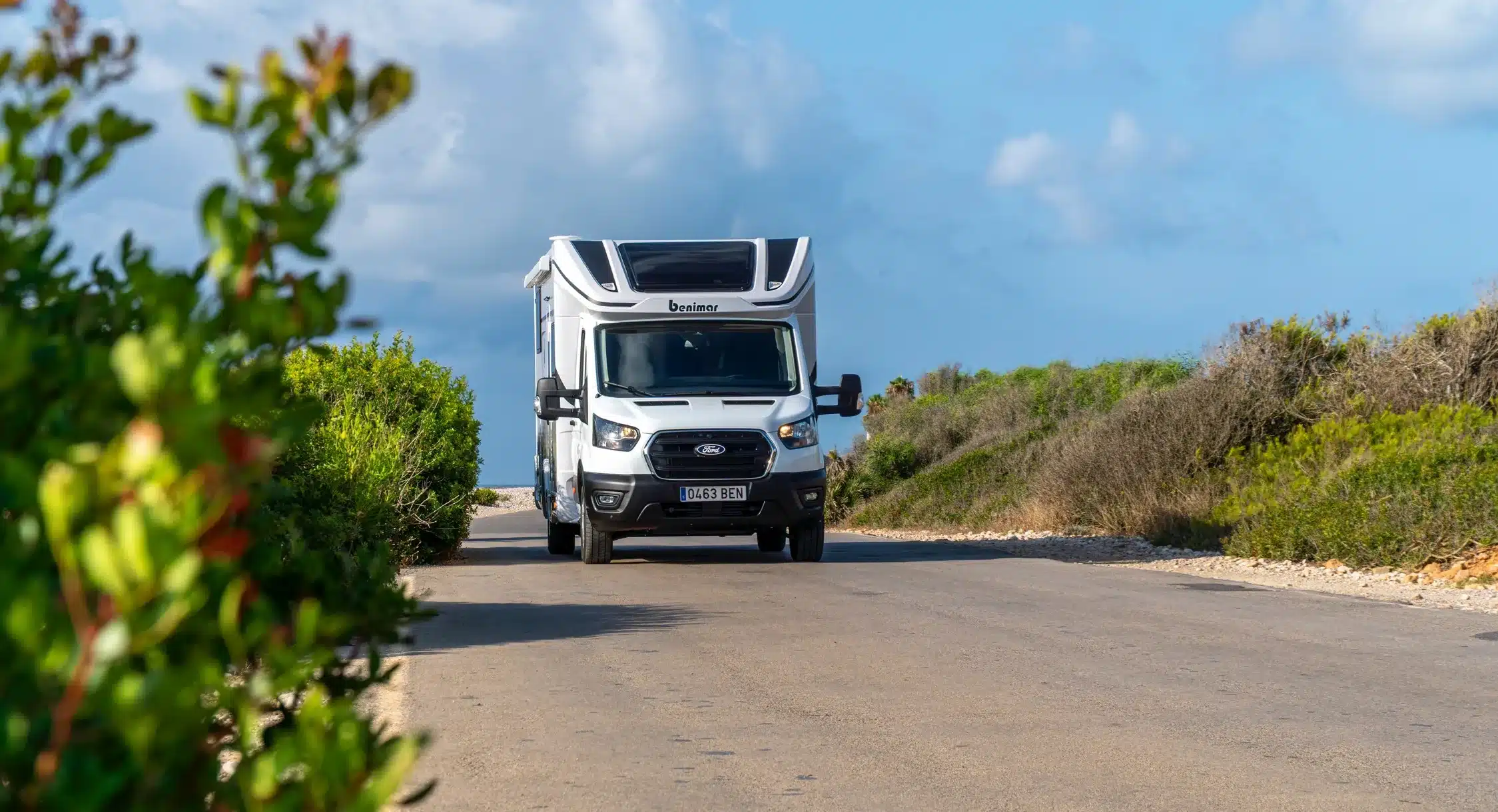 Benimar Tessoro T463 UP Wohnmobil frontal auf ruhiger Straße zwischen Büschen und Himmel.