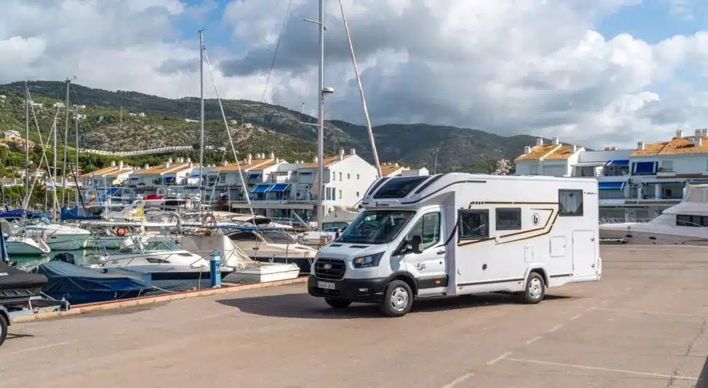Benimar Tessoro T463 UP Wohnmobil vor Yachthafen mit Booten und Bergen im Hintergrund.