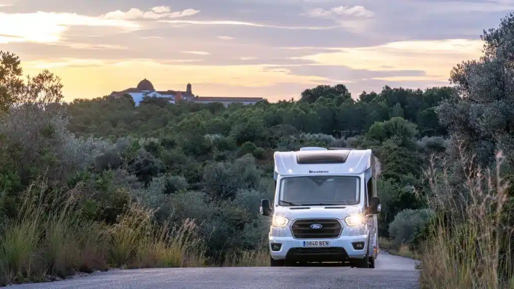 Benimar Y840 Wohnmobil fährt auf einer schmalen Straße durch bewachsene Landschaft, Sonnenuntergang im Hintergrund.