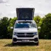 VW T6.1 BIWAK Camper mit geöffnetem Aufstelldach, stehend auf einer grünen Wiese vor Bäumen