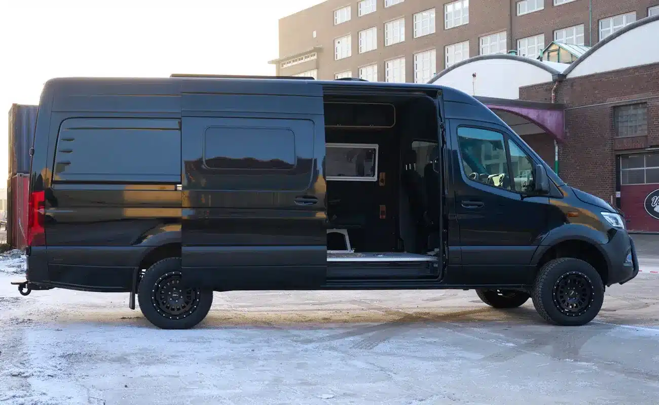 Black Gorilla Campervan Mercedes-Benz Sprinter 4x4 mit offener Seitentür, schwarze Lackierung und Offroad-Ausbau auf Werksgelände.
