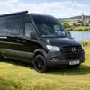 Schwarzer Bresler 694 U Campervan, geparkt auf einer Wiese am See, im Hintergrund ein Dorf mit Kirche.
