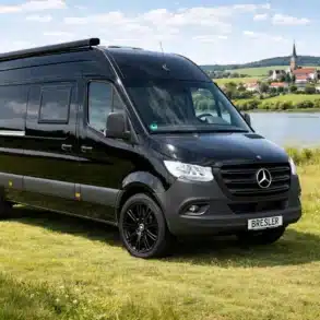 Schwarzer Bresler 694 U Campervan, geparkt auf einer Wiese am See, im Hintergrund ein Dorf mit Kirche.
