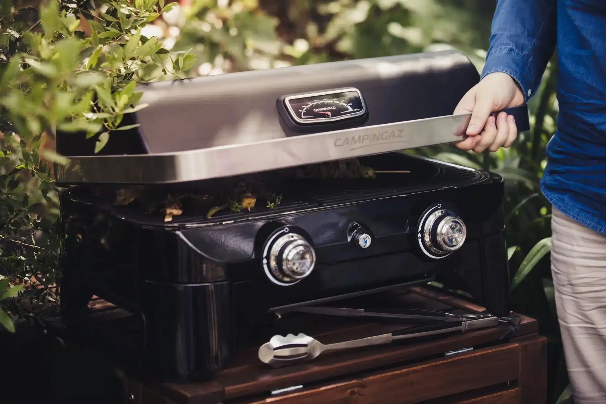 Campingaz Attitude 2100 LX Gas Tischgrill mit geöffnetem Deckel und Grillgut im Grünen