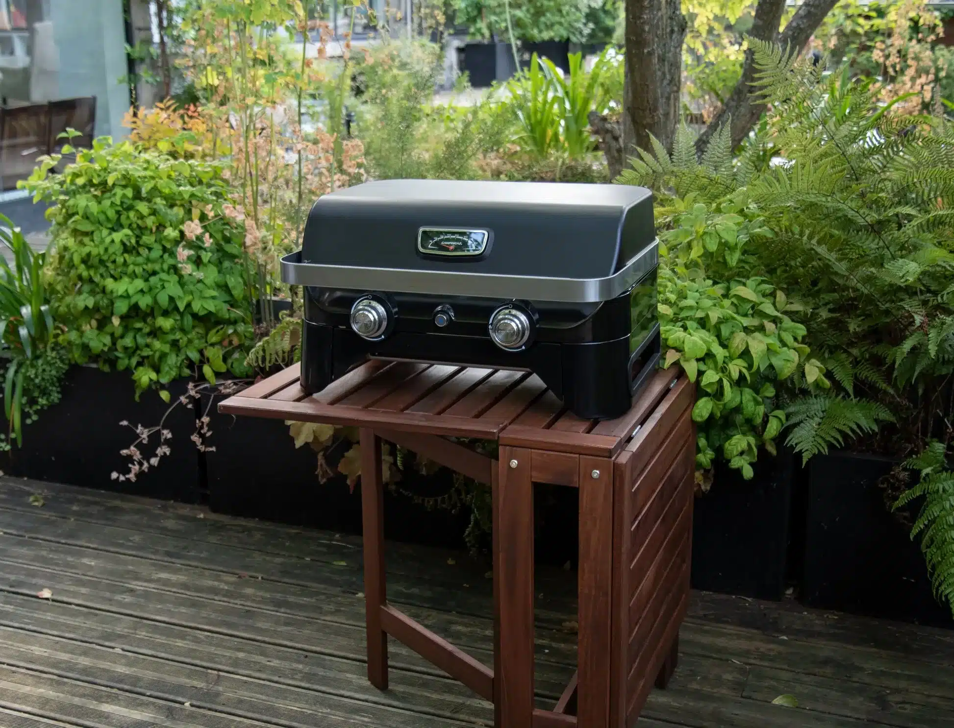 Campingaz Attitude 2100 LX Gas Tischgrill auf Holztisch im Garten, umgeben von Pflanzen