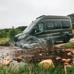 CLIFF 4x4 Greentrek Camper Van fährt durch einen Bach in einer grünen, natürlichen Landschaft.