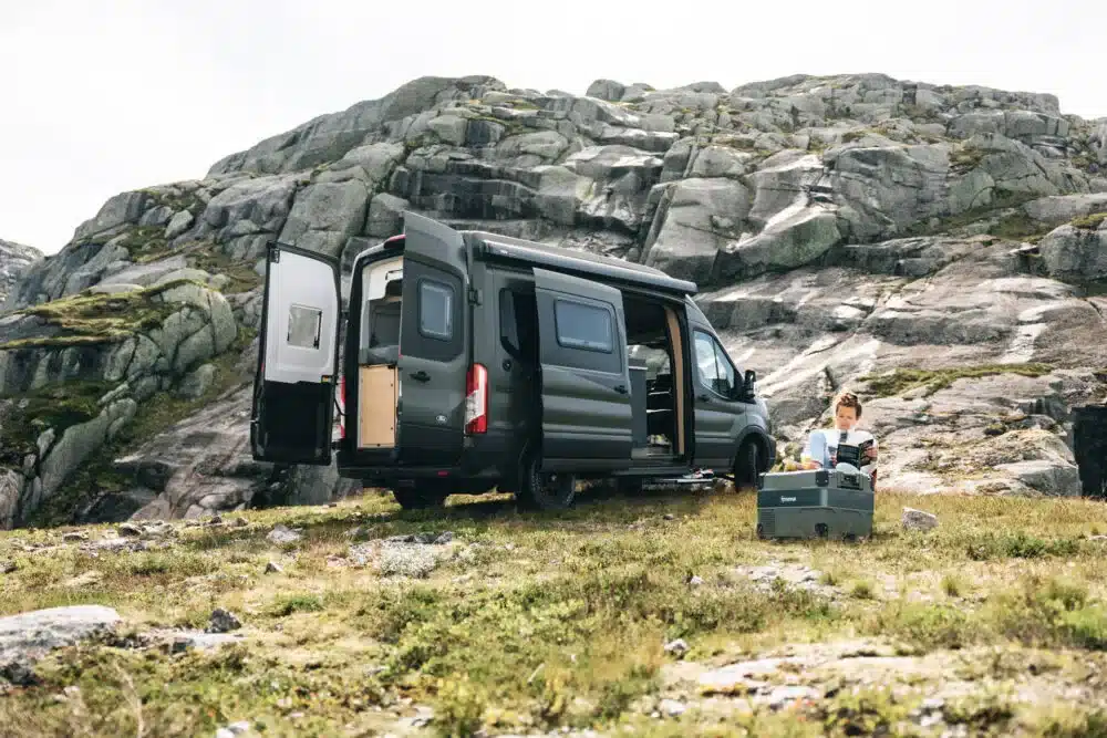 CLIFF 4x4 Greentrek Camper Van steht mit geöffneten Türen in Felslandschaft, daneben Person mit Campingausrüstung.