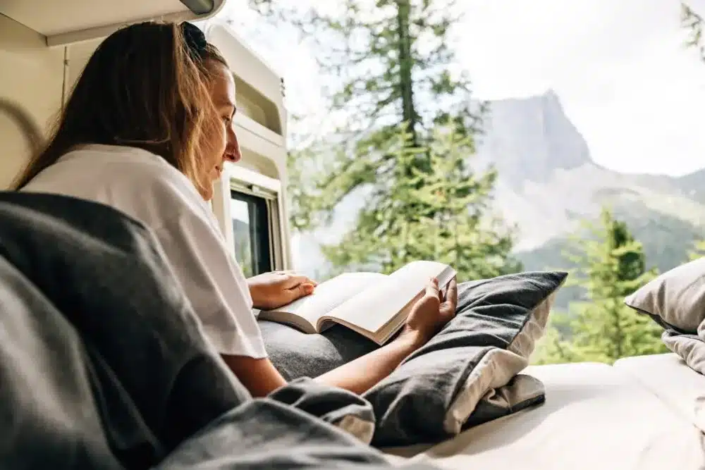 Frau liest im Bett eines Kastenwagens Sunlight CLIFF X mit Ausblick auf Berge durch das Fenster