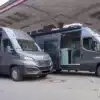 Zwei graue VANME Twins Campervans auf Iveco Daily Basis mit geöffneten Seitentüren und sichtbarem Innenraum an einem Industriegelände.
