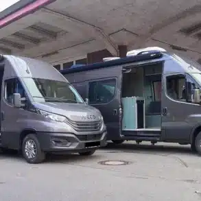 Zwei graue VANME Twins Campervans auf Iveco Daily Basis mit geöffneten Seitentüren und sichtbarem Innenraum an einem Industriegelände.