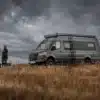 Dovra RIG X-750 Expeditions-Campervan mit Dachträger und Leiter vor bewölktem Himmel in der Natur, zwei Personen und ein Hund gehen neben dem Fahrzeug.
