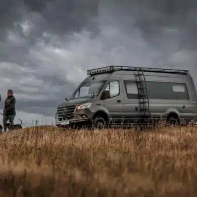Dovra RIG X-750 Expeditions-Campervan mit Dachträger und Leiter vor bewölktem Himmel in der Natur, zwei Personen und ein Hund gehen neben dem Fahrzeug.
