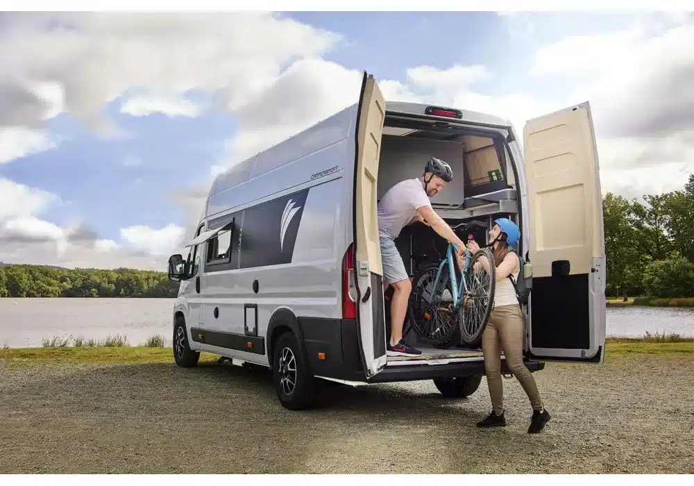 Dreamer Camper Sport mit offenem Heck, zwei Personen verladen ein Fahrrad am Seeufer