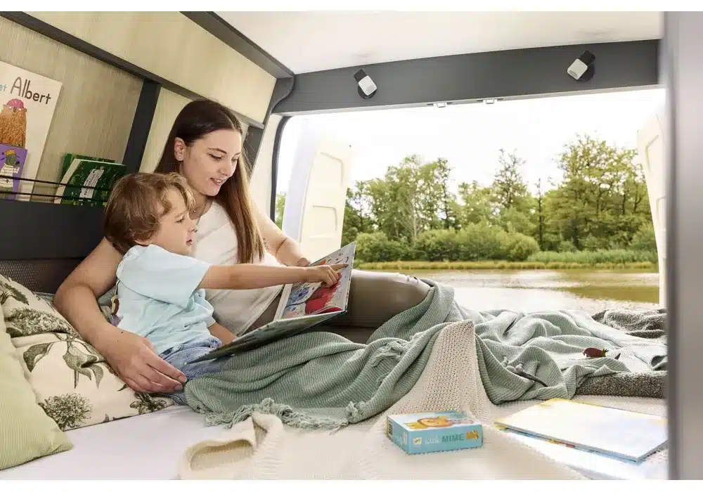 Frau und Kind lesen gemeinsam auf dem Bett im Schlafbereich eines Dreamer Camper Sport mit Blick auf einen Fluss.