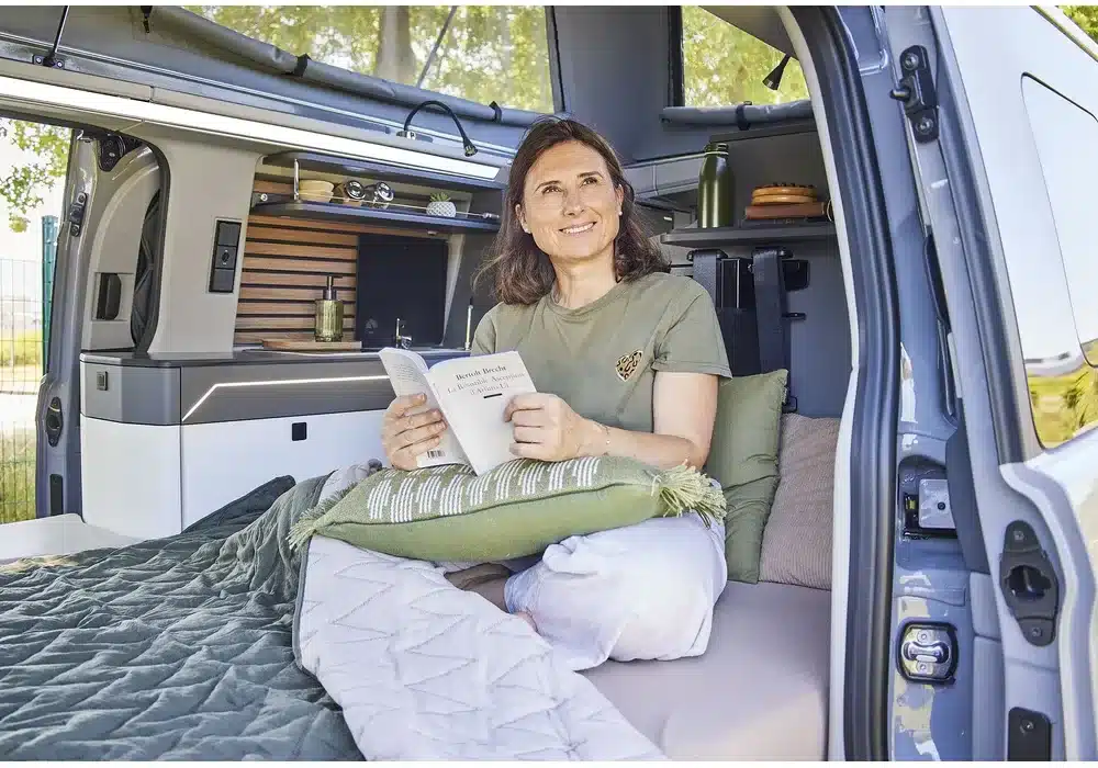 Frau sitzt im Bett des Dreamer Cap Land Camper-Vans und liest, Innenraum mit moderner Ausstattung sichtbar