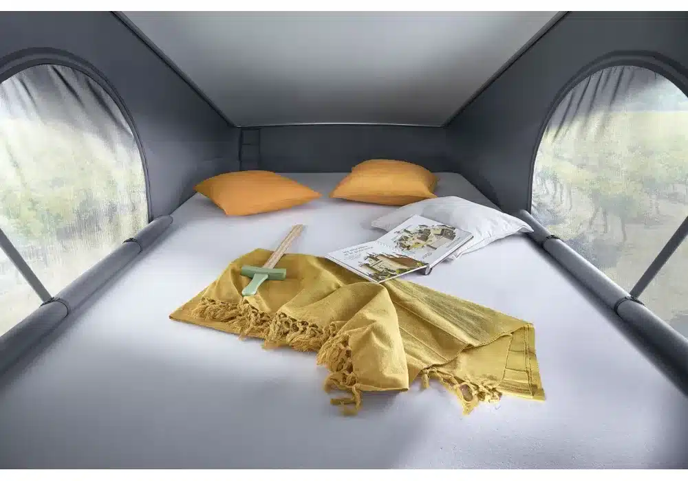 Bett im Aufstelldach des Dreamer D43 UP mit zwei Kissen, einer Decke, einem Buch und Tageslicht durch die seitlichen Fenster.