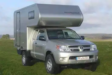 Silberner Toyota Hilux TRAVEL mit GFK-Wohnkabine steht auf einer Wiese