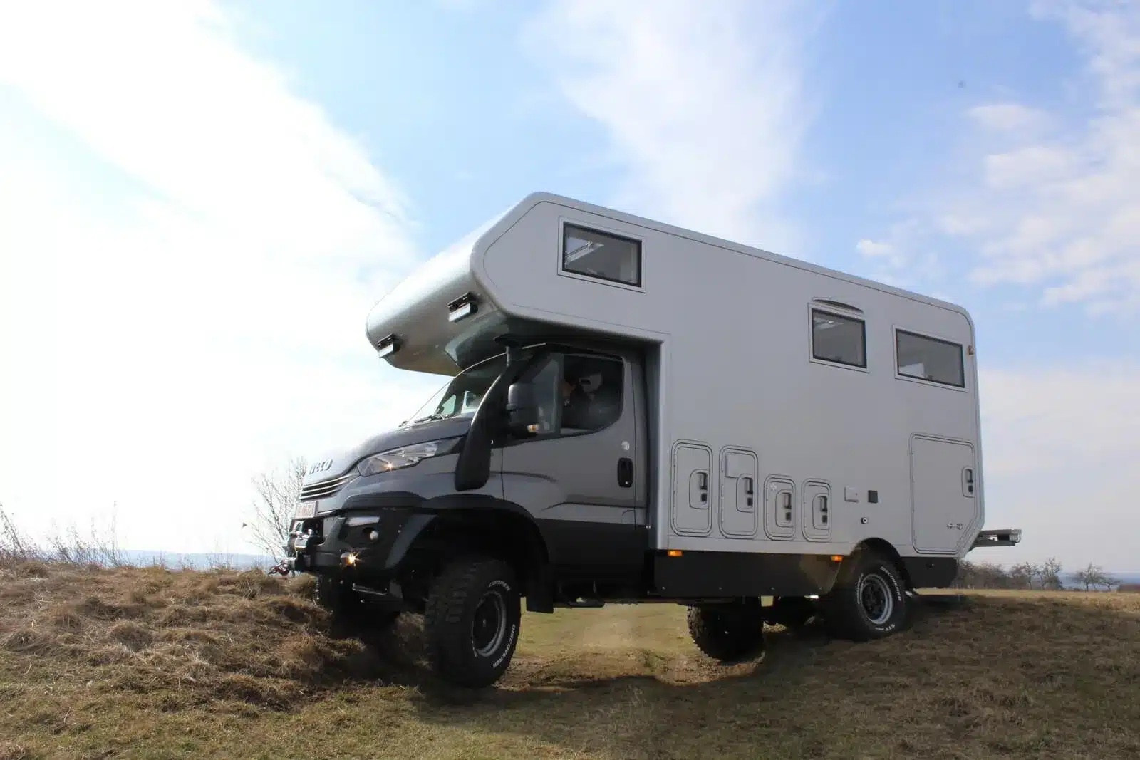 Iveco Daily 4x4 Achleitner mit Woelcke Wohnaufbau fährt über grasbewachsenes, unebenes Gelände.