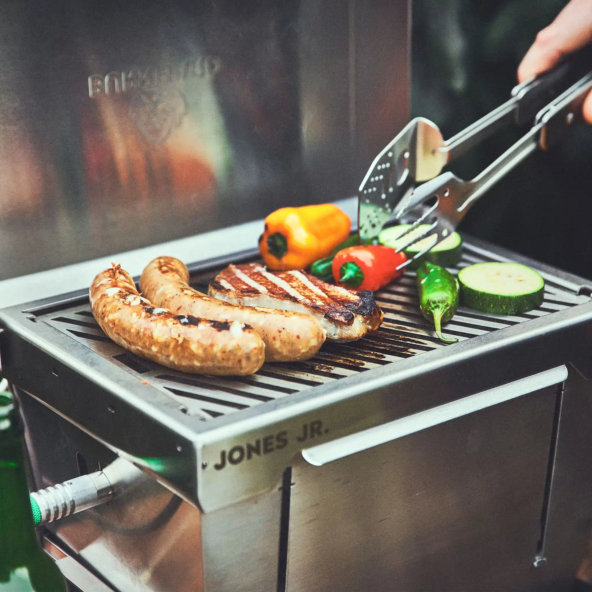 JONES Jr. Campinggrill aus Edelstahl mit Würstchen, Fleisch und Gemüse auf dem Grillrost