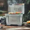 Kompakter Edelstahlgrill JONES Jr. von Burnhard mit Grillgut auf einem Holztisch, angeschlossene Gaskartusche, Outdoor-Einsatz.