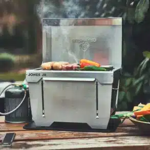 Kompakter Edelstahlgrill JONES Jr. von Burnhard mit Grillgut auf einem Holztisch, angeschlossene Gaskartusche, Outdoor-Einsatz.