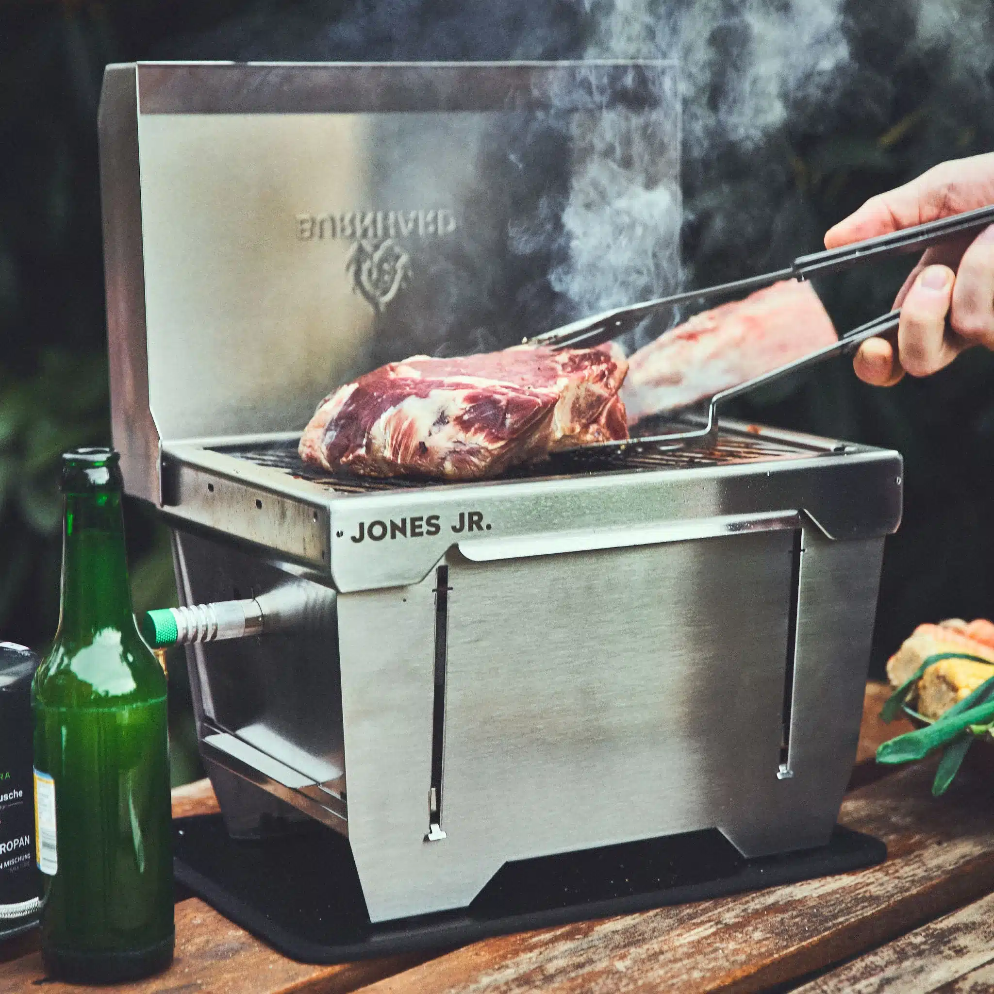 JONES Jr. Campinggrill aus Edelstahl im Einsatz mit aufliegendem Fleisch, Grillzange und Zubehör auf Holztisch.