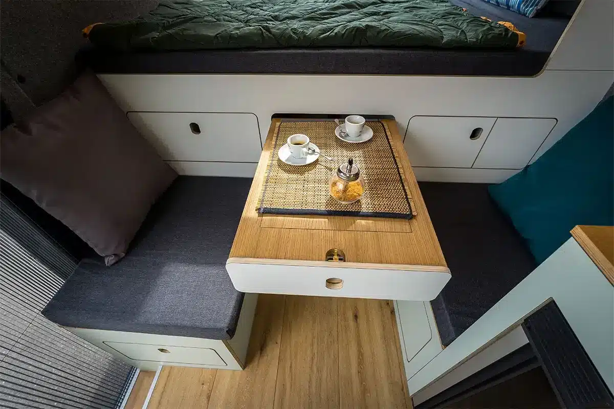 Sitzlounge mit ausziehbarem Tisch und zwei gegenüberliegenden Sitzplätzen im Innenraum des Campervans Libelle, davor ein Querbett.