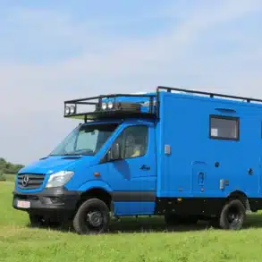 Blauer MB Sprinter 519 CDI 4x4 Expeditionsmobil mit Woelcke-Wohnaufbau, steht auf einer Wiese unter blauem Himmel