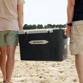 Zwei Personen tragen die Mestic MCCA-42 DC Kompressorkühlbox am Sandstrand neben einem Geländewagen.