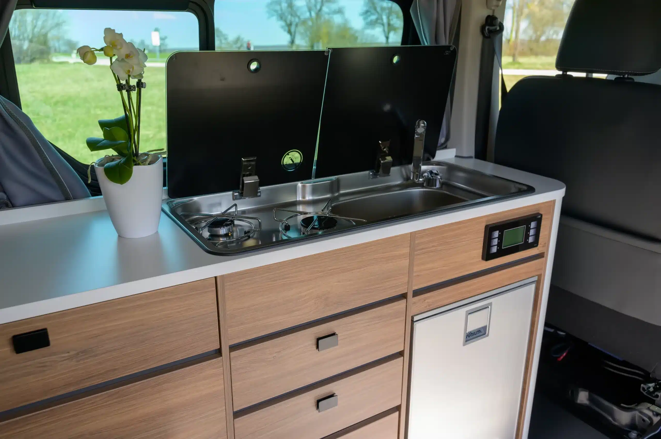 Innenraum eines Campervans mit Küchenzeile, Edelstahlspüle, zweiflammigem Kochfeld und Holzschränken