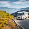 Integriertes Reisemobil MOOVEO INT 78EBFA unterwegs auf einer Straße mit Aussicht auf einen See und grüne Hügellandschaft.