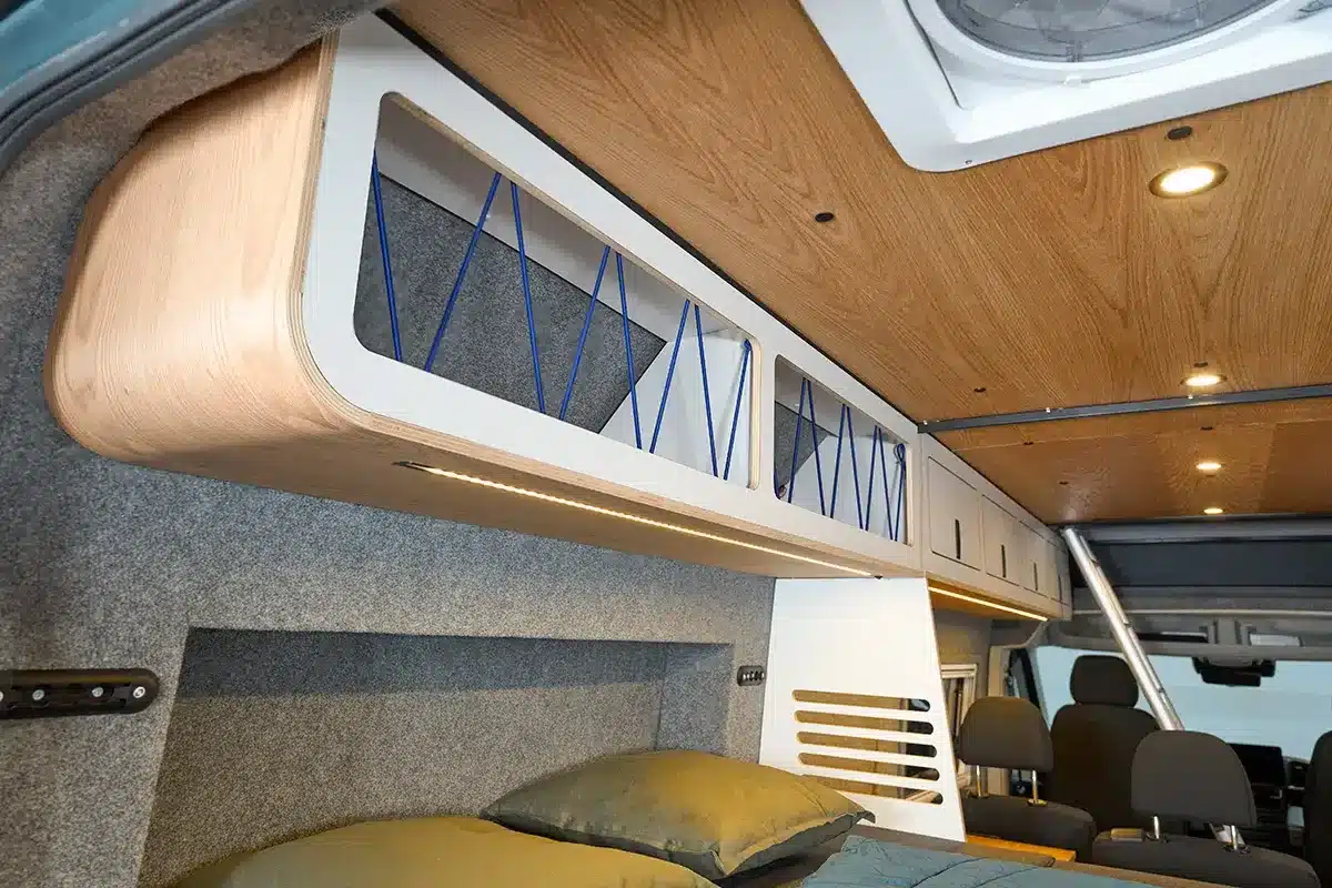 Furnierte Oberschränke mit Abspannseilen und flexiblem Stauraum im Wohnbereich des Multi Racer Campervans, darunter ein Bett und gepolsterte Sitze.