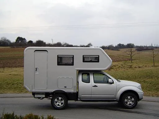 Seitliche Ansicht eines silbernen Nissan Navara 1½ Kabiner mit aufgesetzter GFK-Wohnkabine auf einer Landstraße