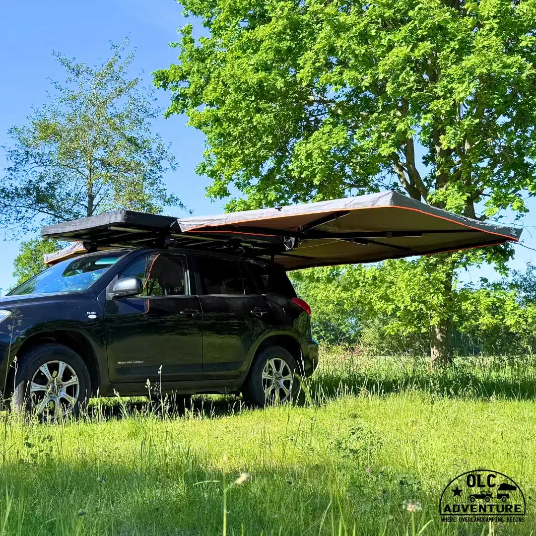 Fahrzeug mit ausgefahrener OLC Adventure 360° Markise auf einer grünen Wiese unter Bäumen, Heckküche im Outdoor-Einsatz.