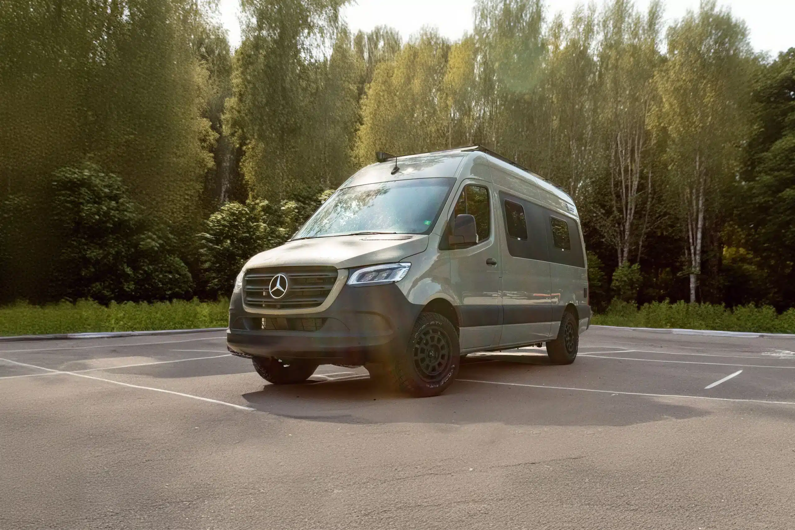 Außenansicht des Pacific Campervans auf Basis eines Mercedes-Benz Sprinter L2H2, stehend auf einem Parkplatz, umgeben von Bäumen.