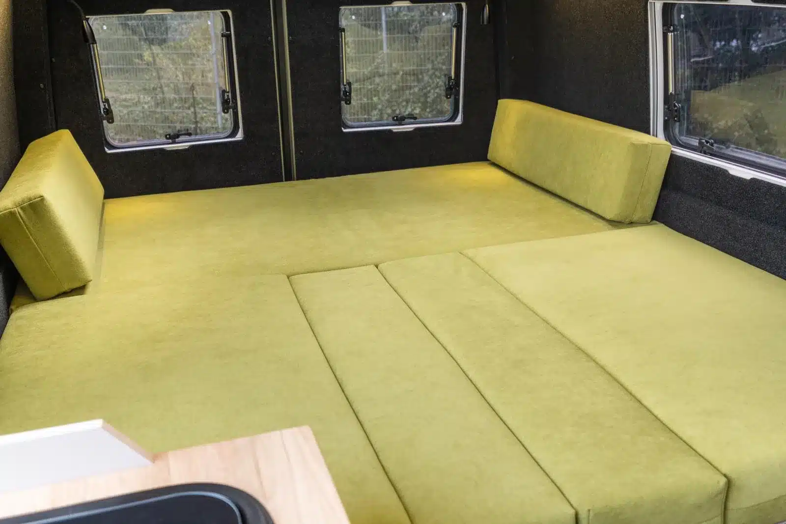 Längsbett mit grünen Kaltschaumpolstern im Innenraum des Pacific Campervan vor zwei Fenstern.