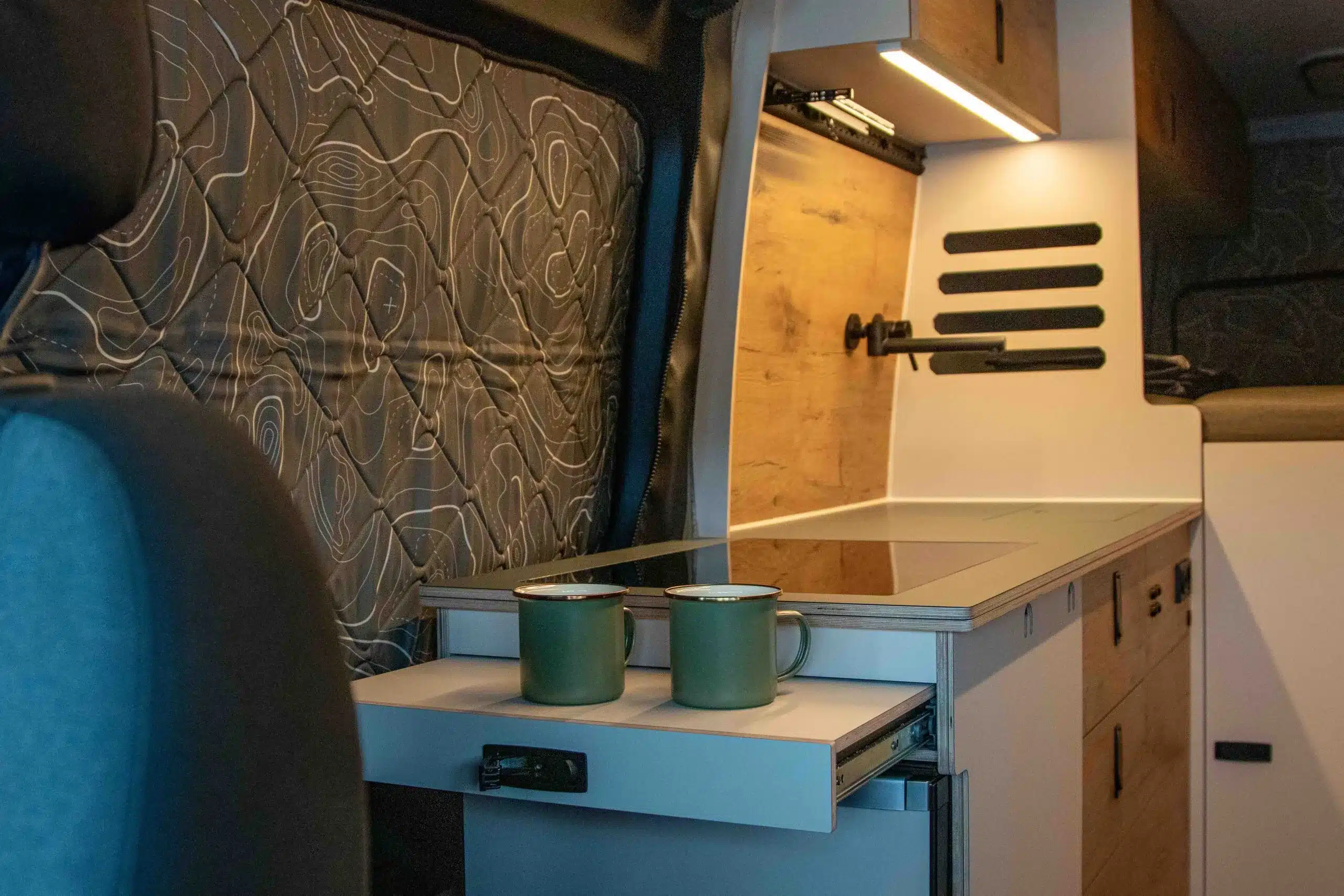 Innenansicht der Camper-Küche im Sky Monster Sprinter mit Induktionskochfeld, Holzfront und zwei Tassen auf dem Auszug.