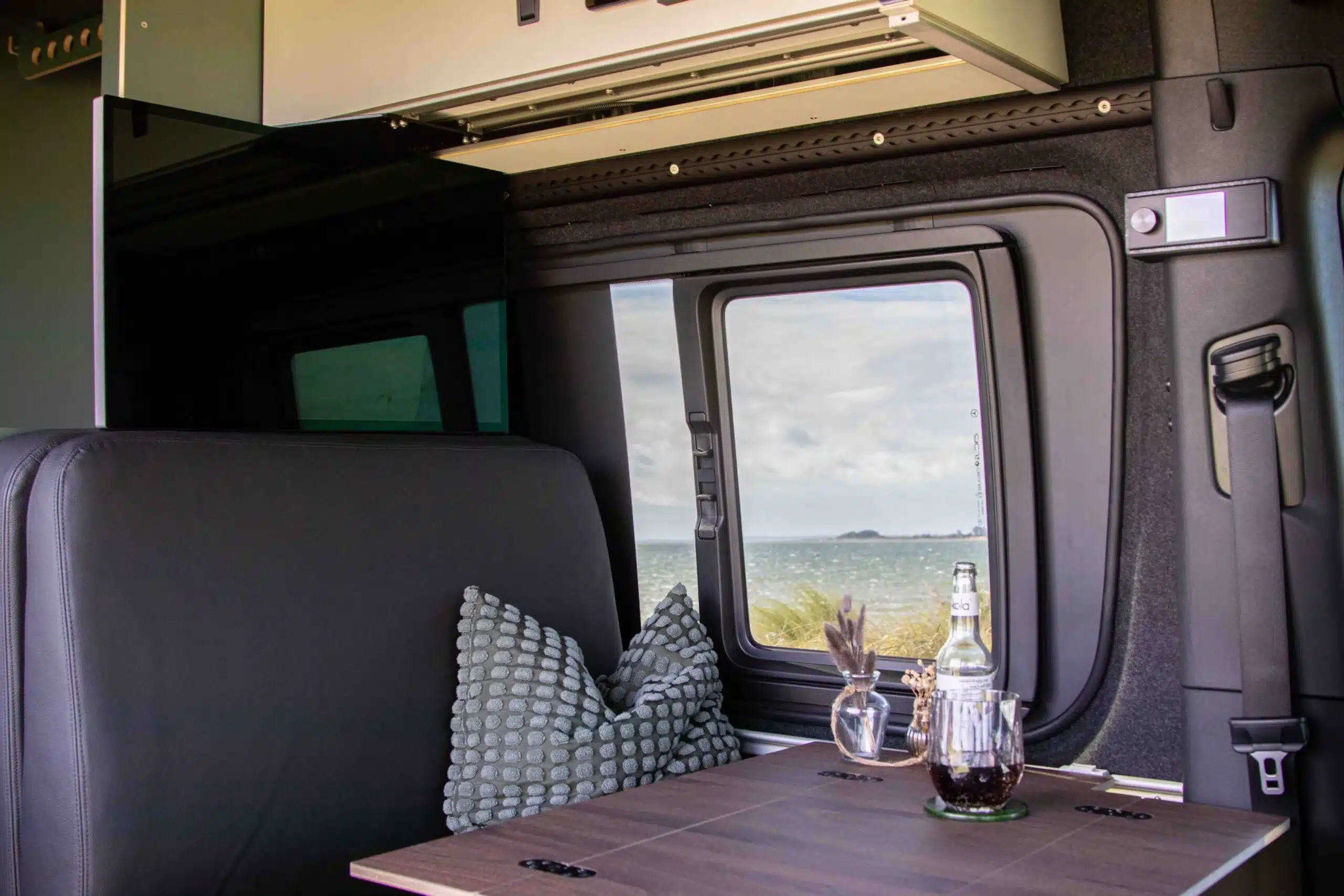 Innenraum eines Offroad-Campers mit Ledersitzbank, Kissen und Holztisch vor großem Fenster mit Ausblick ins Freie.