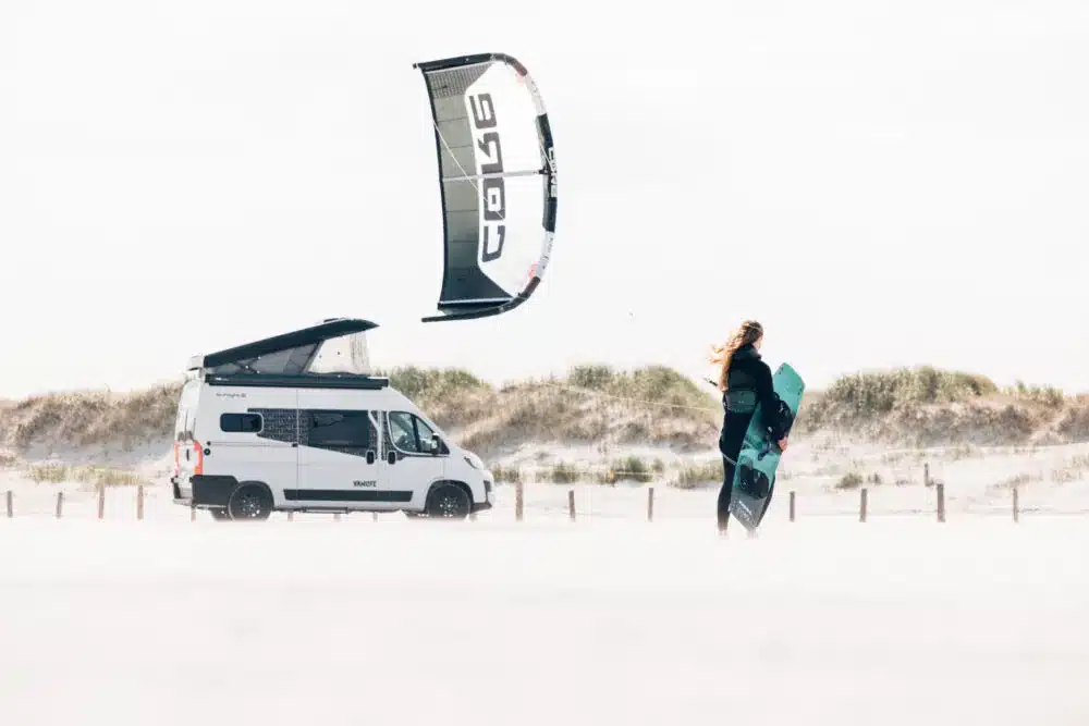 Sunlight VANLIFE Campervan mit Schlafdach am Strand, daneben eine Person mit Kite-Board und Kite im Wind.