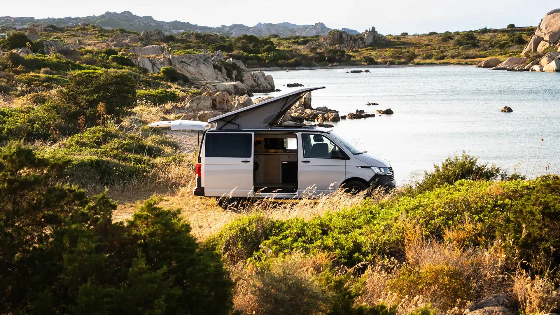 VW T6.1 Expedition Pro Campervan mit geöffnetem Aufstelldach an einem Seeufer inmitten von Felsen und Büschen.