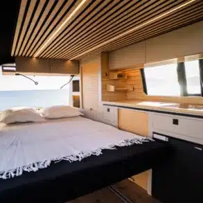 Innenraum eines VW T6.1 Expedition Pro mit ausgeklapptem Bett, Echtholzausbau und Küchenzeile, Blick Richtung Meer.