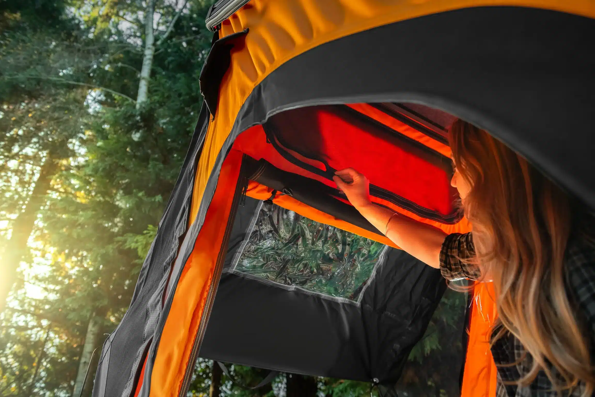 Frau öffnet das Dachfenster eines orange-schwarzen Klappdachzeltes im Wald, Sonnenlicht scheint herein.