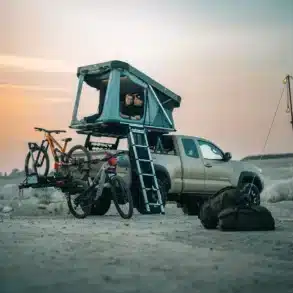 Thule Widesky Dachzelt auf Pick-up, geöffnet, zwei Menschen im Zelt, Fahrräder am Heckträger, draußen auf sandigem Untergrund.