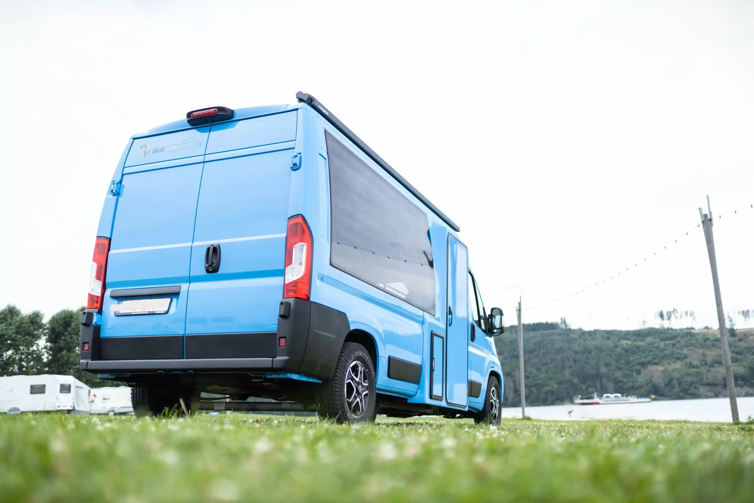 Außenansicht des TS BusCamp TIPI Campingbusses auf Basis Fiat Ducato, himmelblau lackiert, geparkt auf einer Wiese am Wasser.