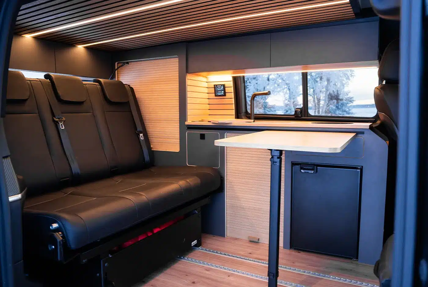 Innenraum eines Mercedes V-Klasse Expedition Campervans mit drei Sitzplätzen, Tisch, Küchenzeile und moderner Holzverkleidung.