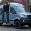 Mercedes Benz Sprinter Campervan Wandertaube mit geöffneten Türen und sichtbarem Innenausbau, abgestellt vor Industriegebäude.