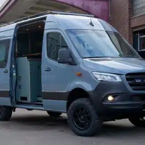Mercedes Benz Sprinter Campervan Wandertaube mit geöffneten Türen und sichtbarem Innenausbau, abgestellt vor Industriegebäude.
