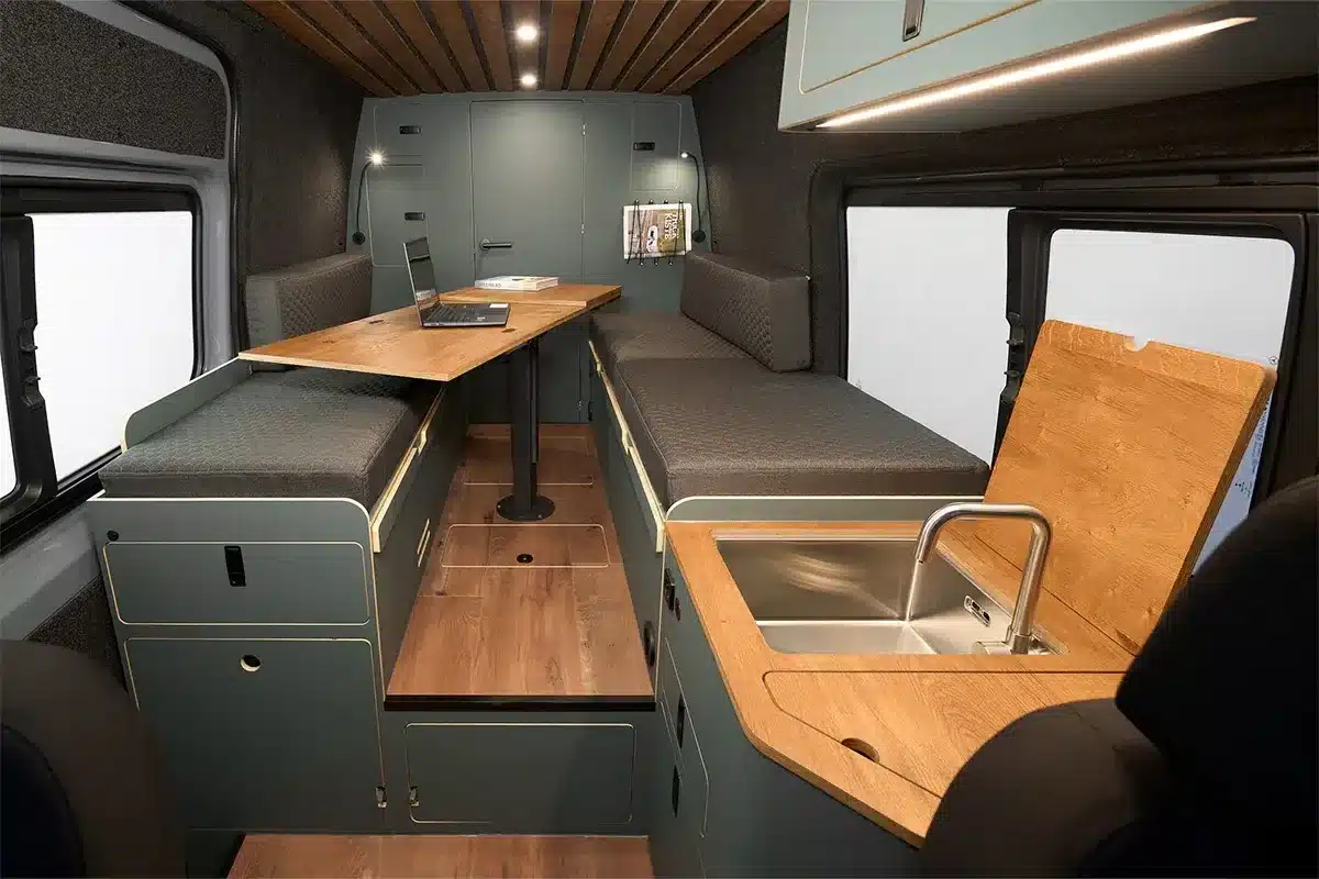 Innenraum eines ausgebauten Mercedes Sprinter Campervans mit Sitzbank, Arbeitsplatz und Küchenbereich mit Spüle.