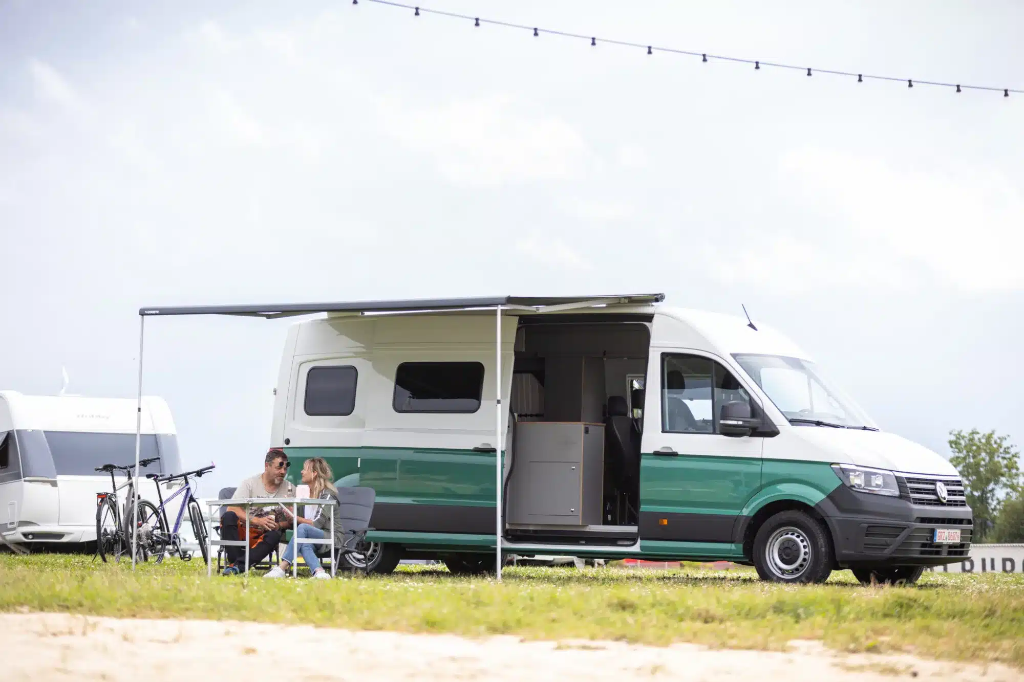 WIGWAM Campervan auf VW Crafter Basis mit ausgefahrener Markise, Paar sitzt davor an einem Tisch mit Fahrrädern im Hintergrund.